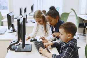 Crianças aprendem juntas em um laboratório de informática escolar, utilizando o computador como ferramenta de educação digital em ambiente colaborativo e inclusivo.