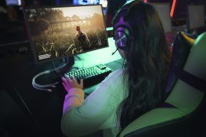 Mulher com headset jogando em um computador em ambiente de lan house com iluminação neon, representando o crescente uso de plataformas de streaming nacional para games e entretenimento digital no Brasil.