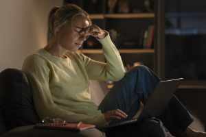 Mulher jovem sentada no sofá à noite, usando notebook e demonstrando sinais de cansaço e estresse, representando o impacto psicológico da tecnologia no dia a dia.