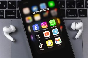 Smartphone exibindo uma pasta de aplicativos de redes sociais, incluindo Facebook, Instagram, WhatsApp, X, YouTube, Telegram, TikTok, Snapchat e Gmail, posicionado sobre um teclado de notebook com fones de ouvido brancos ao lado.